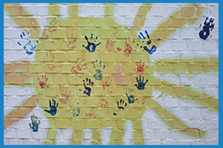 White brick wall with a large yellow sun painted over, with childrens handprints