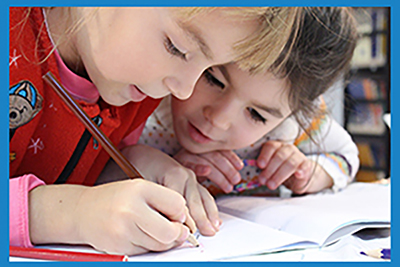 two children writing together, leaning over a book